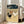 May include: Vintage-style artwork print showing two roosters - one black with red comb and one white - standing on sandy ground beneath a dramatic cloudy sky. The illustration has a textured, grainy appearance reminiscent of mid-century printmaking. Displayed in a light wood frame leaning against a white wall next to a beige upholstered chair in a bright, minimalist living room with large windows.