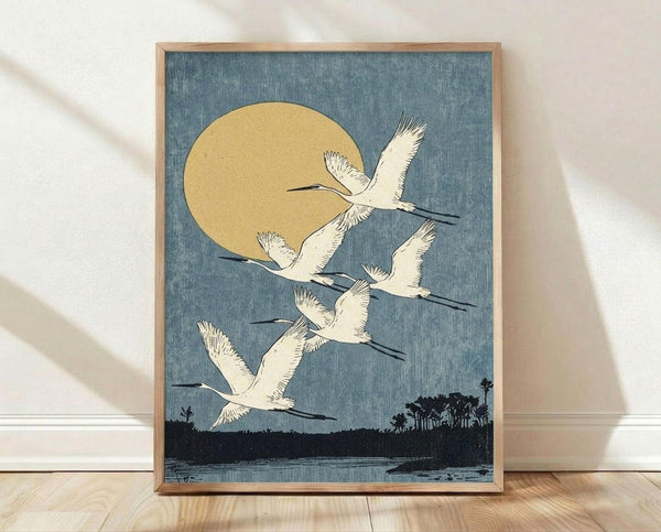 May include: Framed vintage Japanese woodblock print featuring five white cranes flying against a dusty blue sky with golden sun. Dark silhouetted landscape with trees and water below. Natural wood frame leaning against white wall on light wooden floor with soft shadows.