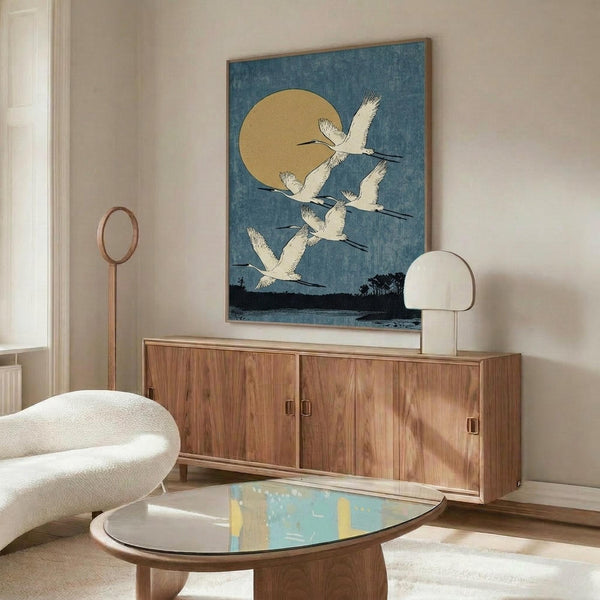 May include: Japanese woodblock print artwork displayed above a wooden sideboard in a modern living room. The artwork features white cranes flying across a blue sky with a golden moon, framed in natural wood. Room includes cream walls, white curved chair, oval glass coffee table, and minimalist decor with warm wood tones throughout.