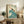 May include: Vintage-style travel poster featuring swimmer diving into stylized blue-green wave, cream background with teal text reading "The Seaside Calls" and "Go By Train Take a Kodak." Framed in light wood, displayed on rustic wooden sideboard with potted fern, books, ceramic vase, and cream upholstered chair visible in cozy living room setting.