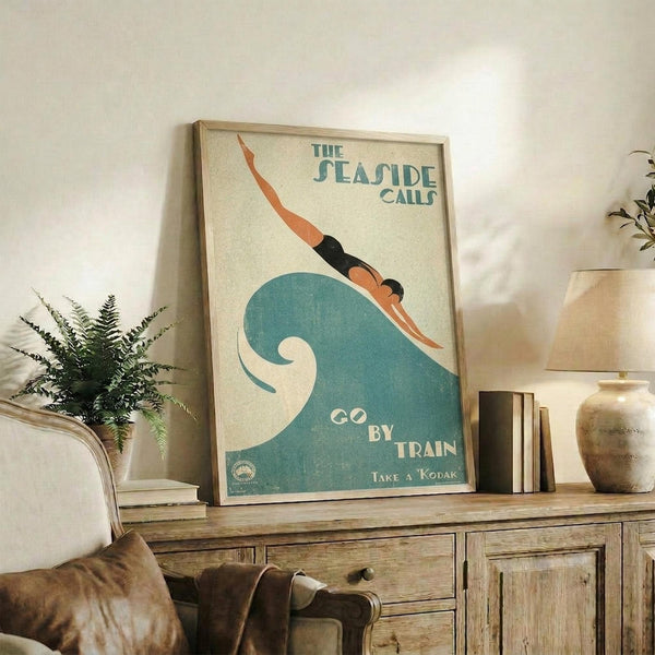 May include: Vintage-style travel poster featuring swimmer diving into stylized blue-green wave, cream background with teal text reading "The Seaside Calls" and "Go By Train Take a Kodak." Framed in light wood, displayed on rustic wooden sideboard with potted fern, books, ceramic vase, and cream upholstered chair visible in cozy living room setting.