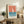 May include: Framed vintage-style poster with coral and blue sailboat design displayed on rustic wooden sideboard next to potted fern, ceramic vases, and books in cozy living room setting with beige chair and neutral walls