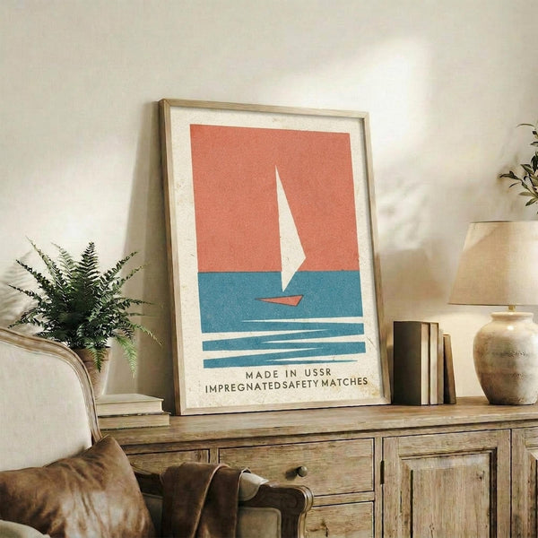 May include: Framed vintage-style poster with coral and blue sailboat design displayed on rustic wooden sideboard next to potted fern, ceramic vases, and books in cozy living room setting with beige chair and neutral walls
