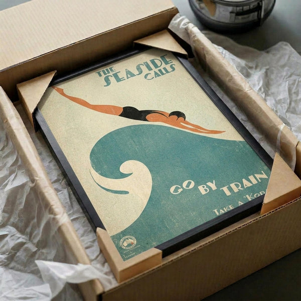 May include: Vintage-style poster showing a swimmer diving over stylized teal waves with "THE SEASIDE CALLS" and "GO BY TRAIN" text, displayed in black frame inside cardboard shipping box with protective tissue paper wrapping.