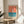 May include: Framed vintage-style poster with coral orange sky, white sailboat, and turquoise blue water with horizontal wave lines. Text reads "Made in USSR Impregnated Safety Matches" at bottom. Light wood frame sits on round wooden side table next to beige upholstered chair with white pillow. Green plant leaves visible on left, warm natural sunlight streaming across white walls creating soft shadows.