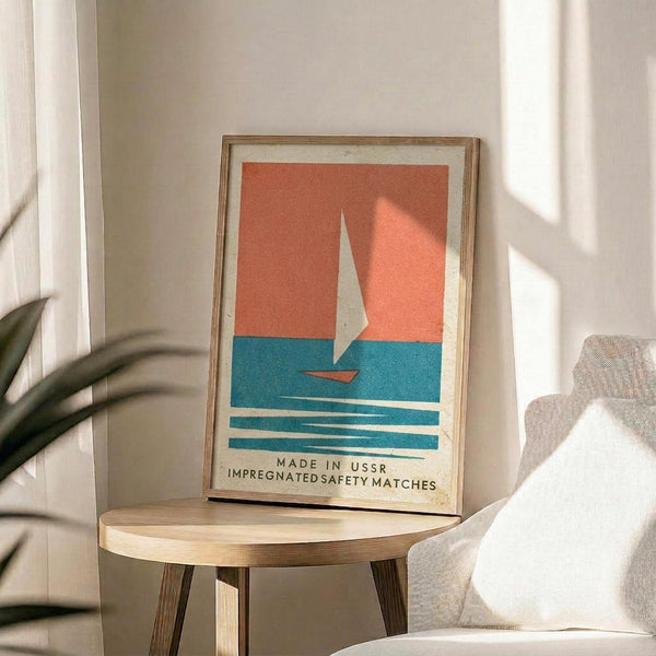 May include: Framed vintage-style poster with coral orange sky, white sailboat, and turquoise blue water with horizontal wave lines. Text reads "Made in USSR Impregnated Safety Matches" at bottom. Light wood frame sits on round wooden side table next to beige upholstered chair with white pillow. Green plant leaves visible on left, warm natural sunlight streaming across white walls creating soft shadows.