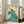 May include: Vintage-style travel poster displayed in bright living room with white walls and beige sofa. Artwork features art deco design with turquoise wave, orange figure diving, cream background, and text reading "The Seaside Calls" and "Go By Train Take a Kodak". Poster has light wood frame and leans against wall on hardwood floor near large windows.