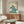 May include: Vintage travel poster featuring swimmer diving into turquoise wave, displayed in wooden frame above walnut credenza in modern living room. Cream-colored spherical chair and brass floor lamp visible alongside oval glass coffee table. Poster shows art deco style illustration with beige background and blue-green wave design.