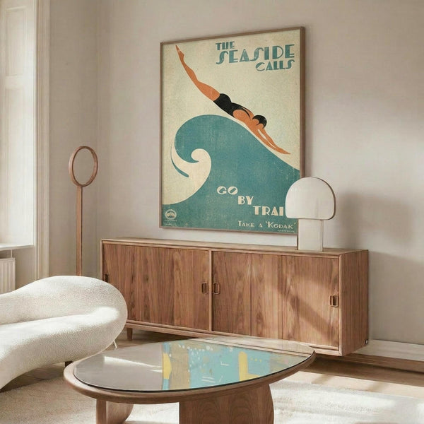 May include: Vintage travel poster featuring swimmer diving into turquoise wave, displayed in wooden frame above walnut credenza in modern living room. Cream-colored spherical chair and brass floor lamp visible alongside oval glass coffee table. Poster shows art deco style illustration with beige background and blue-green wave design.