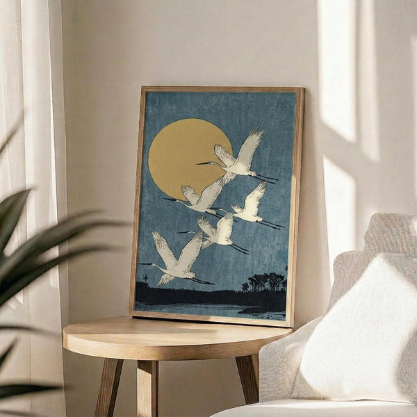 May include: Vintage Japanese woodblock print featuring five white cranes flying against a blue textured sky with golden moon and dark landscape silhouette below. Art displayed in thin wooden frame on round wooden side table next to beige upholstered chair with white pillow. Natural sunlight streams across neutral-toned room with plant leaves visible in corner.