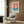 May include: Vintage-style poster featuring a white sailboat on blue water against coral sky, framed in light wood and displayed on dark wood paneled wall above beige upholstered chair with geometric pillow. Modern reading nook with brass floor lamp and side table with open book. The poster shows minimalist nautical design with text at bottom in cream and coral color palette.