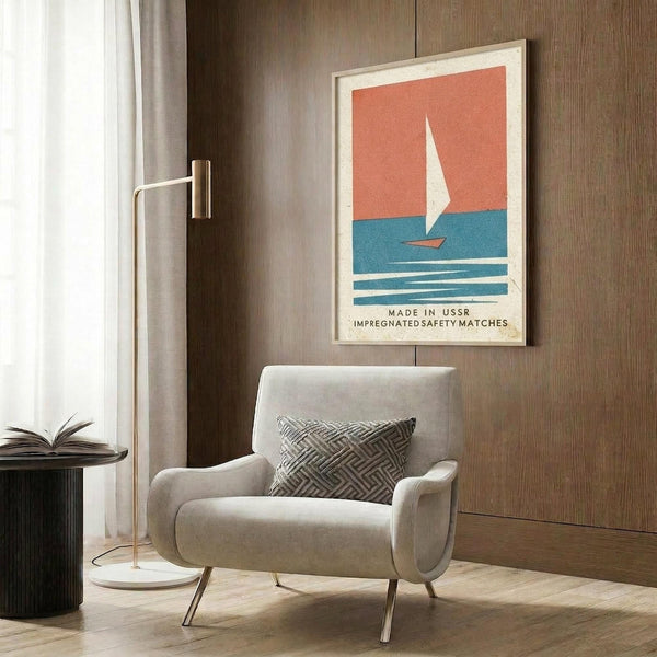 May include: Vintage-style poster featuring a white sailboat on blue water against coral sky, framed in light wood and displayed on dark wood paneled wall above beige upholstered chair with geometric pillow. Modern reading nook with brass floor lamp and side table with open book. The poster shows minimalist nautical design with text at bottom in cream and coral color palette.