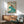 May include: Vintage-style travel poster featuring a stylized swimmer diving through a large teal wave, with text "THE SEASIDE CALLS" and "GO BY TRAIN - TAKE A KODAK" in art deco typography. The framed print hangs above a wooden sideboard with white ceramic lamp, next to a mid-century modern teal upholstered armchair with wooden frame. Warm sunlight streams across white walls creating geometric shadows.