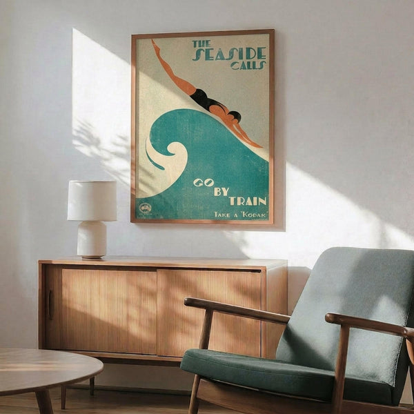 May include: Vintage-style travel poster featuring a stylized swimmer diving through a large teal wave, with text "THE SEASIDE CALLS" and "GO BY TRAIN - TAKE A KODAK" in art deco typography. The framed print hangs above a wooden sideboard with white ceramic lamp, next to a mid-century modern teal upholstered armchair with wooden frame. Warm sunlight streams across white walls creating geometric shadows.