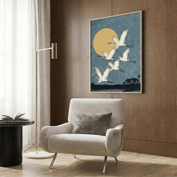 May include: A framed vintage Japanese woodblock print featuring white cranes flying against a blue sky with golden moon, displayed on wood-paneled wall above a cream upholstered armchair with geometric pillow. Modern floor lamp and round side table with open book complete the elegant reading nook scene.