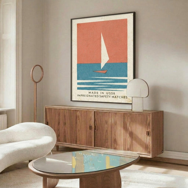 May include: Vintage-style poster with coral pink sky, blue water, and white sailboat hung above a walnut credenza in modern living room. Black thin frame, white matting. Room features cream walls, white curved chair, oval glass coffee table, brass floor lamp, and natural lighting from window.