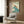 May include: Vintage-style poster featuring a swimmer diving over a stylized teal wave, with text reading "The Seaside Calls" and "Go By Train" with "Take a Kodak" at bottom. Displayed in light wood frame on warm wood-paneled wall above modern cream armchair with geometric patterned pillow. Gold floor lamp and dark side table with open book complete the cozy reading nook setting.