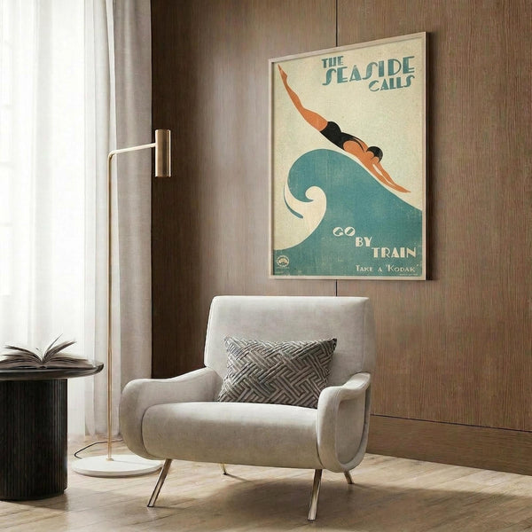May include: Vintage-style poster featuring a swimmer diving over a stylized teal wave, with text reading "The Seaside Calls" and "Go By Train" with "Take a Kodak" at bottom. Displayed in light wood frame on warm wood-paneled wall above modern cream armchair with geometric patterned pillow. Gold floor lamp and dark side table with open book complete the cozy reading nook setting.