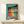 May include: Framed vintage-style poster with turquoise background showing five women in colorful dresses (red, orange, yellow, green, blue) dancing in synchronized pose with arms raised. Text reads "Dance More" in orange lettering. Wood frame sits on light wooden shelf next to small wrapped white gift box against cream wall.