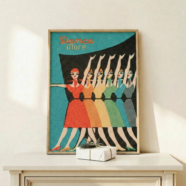 May include: Framed vintage-style poster with turquoise background showing five women in colorful dresses (red, orange, yellow, green, blue) dancing in synchronized pose with arms raised. Text reads "Dance More" in orange lettering. Wood frame sits on light wooden shelf next to small wrapped white gift box against cream wall.