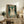 May include: Framed vintage-style poster featuring a tabby cat wearing black headphones on teal background with cream text and musical notes, displayed on rustic wooden sideboard alongside potted fern, ceramic vase, books, and brown leather armchair in cozy living room setting with white walls