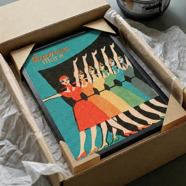 May include: Framed vintage-style poster with turquoise background showing retro illustration of dancing women in colorful dresses - one in red, others in yellow and green - with raised arms in synchronized dance pose. Text reads "Dance More" in orange lettering. Black frame sits in cardboard shipping box with white tissue paper packaging on wooden surface.