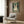 May include: Framed poster on wood-paneled wall showing tabby cat wearing black headphones with text about music and murder in vintage-style design with green background. Modern beige armchair with geometric pillow, gold floor lamp, and side table with open book in contemporary room setting with neutral tones.