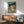 May include: Mid-century modern living room with teal and wood furniture featuring a framed vintage-style poster showing five dancing women in colorful dresses - red, orange, yellow, green, and blue - with "Dance More" text on turquoise background, displayed above wooden sideboard next to white ceramic lamp and sage green upholstered chair with wooden arms.