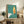 May include: Mid-century modern poster with turquoise textured background featuring a yellow and orange flame design above white geometric shapes, displayed in light wood frame on rustic wooden sideboard alongside fern plant, books, ceramic vase, and beige armchair in cozy living room setting with neutral walls