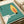 May include: Vintage Polish matchbox label artwork displayed on white paper with cream ribbon nearby. Features a stylized leaf design in yellow and orange on teal speckled background, with white hand silhouette below. Text appears in Polish along edges. Retro mid-century modern aesthetic with grainy textured printing style typical of 1960s Eastern European design.