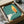 May include: Framed vintage poster print in cardboard box with protective packaging. Green textured background features minimalist illustration of white hand holding lit match with yellow and orange flame. Polish text visible at bottom. Print appears square format in black frame, nestled in brown shipping box with tissue paper wrapping.