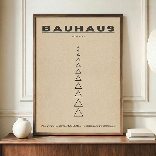 May include: Framed beige poster with black text reading BAUHAUS and "LESS IS MORE" above a vertical row of outlined triangles decreasing in size, displayed on wooden sideboard next to white ceramic vase and books against cream paneled wall in modern interior setting.
