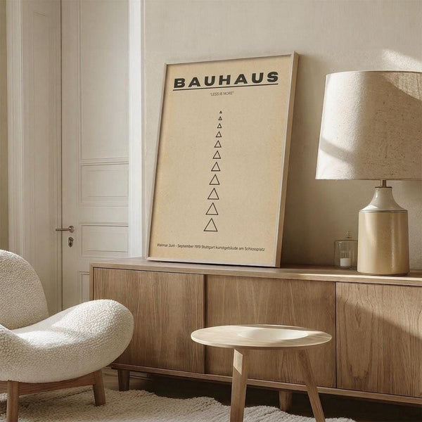 a minimalist living room with a large framed poster of Bauhaus art hanging on the wall, a white armchair, a wooden sideboard, and a round wooden table.