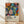May include: Colorful abstract botanical poster in wooden frame leaning against white wall on light wood floor. Artwork features vibrant flowers, leaves, and circular shapes in orange, blue, pink, yellow, and green against black polka dot background. Text at bottom reads "YAYOI KUSAMA" with Japanese characters and museum information. Soft natural lighting creates diagonal shadows across the scene.