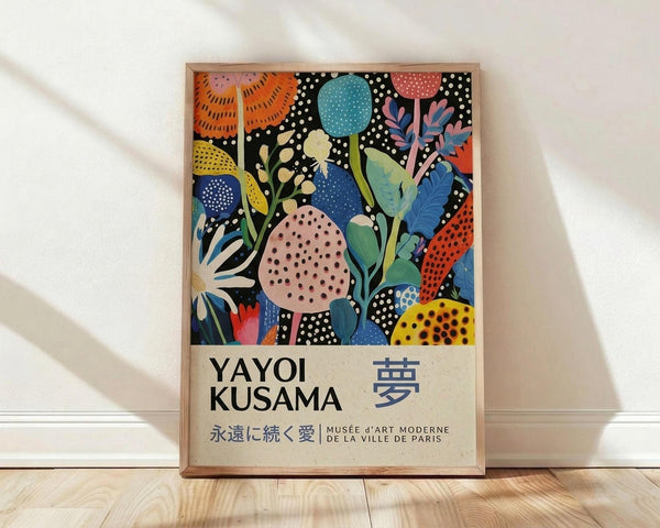 May include: Colorful abstract botanical poster in wooden frame leaning against white wall on light wood floor. Artwork features vibrant flowers, leaves, and circular shapes in orange, blue, pink, yellow, and green against black polka dot background. Text at bottom reads "YAYOI KUSAMA" with Japanese characters and museum information. Soft natural lighting creates diagonal shadows across the scene.