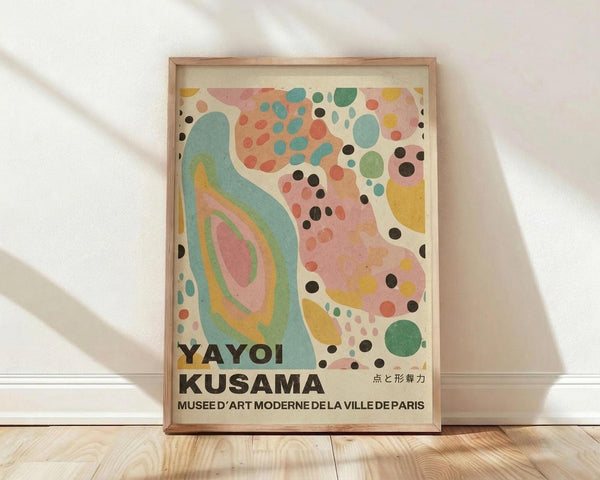 May include: Abstract art poster with colorful organic shapes and polka dots in a light wood frame, leaning against white wall on wooden floor. Features flowing turquoise forms, scattered circles in pink, yellow, green, and black dots throughout. Text reads "YAYOI KUSAMA" and "MUSEE D'ART MODERNE DE LA VILLE DE PARIS" in black letters at bottom on cream background.