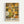 Framed art print featuring a dense pattern of stylized flowers and polka dots in warm autumn colors including yellows, oranges, greens, browns, and cream. The abstract floral design fills the entire composition with overlapping daisy-like blooms of varying sizes interspersed with circular dots and geometric patterns. Text at bottom reads "YAYOI KUSAMA" with Japanese characters and "NAGOYA" in a simple font. Displayed in a light wood frame against a white wall.