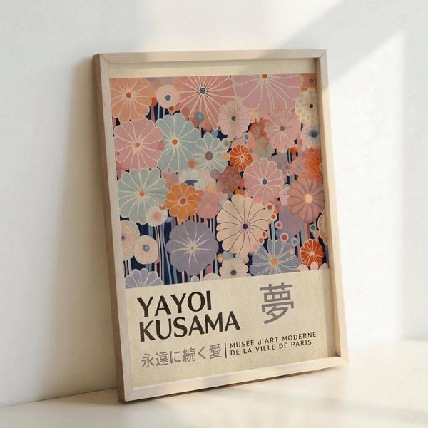 Framed poster featuring a colorful floral pattern with stylized daisy-like flowers in soft pastels including pink, sage green, orange, and lavender against a navy blue background. The artwork shows "YAYOI KUSAMA" in bold black letters with Japanese characters and "MUSÉE d'ART MODERNE DE LA VILLE DE PARIS" below. The light wood frame leans against a white wall, casting a soft shadow.