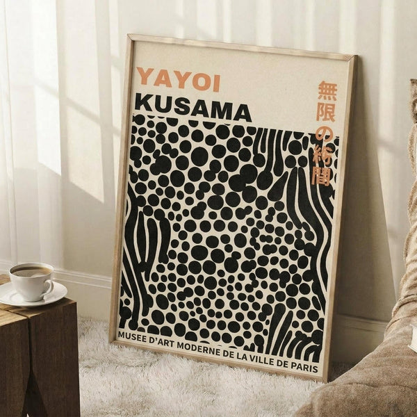 May include: Framed poster with cream background featuring orange "YAYOI KUSAMA" text and black polka dot pattern with flowing wavy lines, displayed leaning against white paneled wall next to wooden side table with white coffee cup and saucer, on textured cream area rug beside beige upholstered furniture in bright natural lighting.