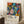 May include: Framed colorful botanical art poster leaning against white wall corner next to beige armchair and wooden side table with white coffee cup. Artwork features vibrant abstract plants, flowers and leaves in orange, blue, pink, yellow with polka dot patterns on black background. Text shows artist name and Japanese characters with Paris museum reference at bottom.