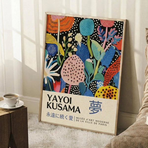 May include: Framed colorful botanical art poster leaning against white wall corner next to beige armchair and wooden side table with white coffee cup. Artwork features vibrant abstract plants, flowers and leaves in orange, blue, pink, yellow with polka dot patterns on black background. Text shows artist name and Japanese characters with Paris museum reference at bottom.