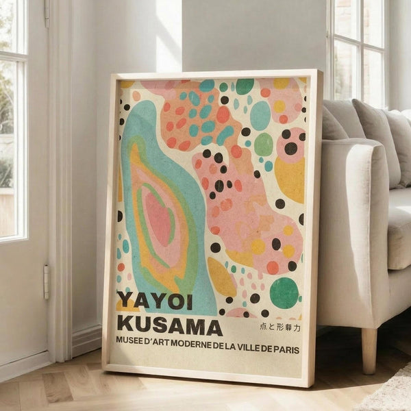 May include: Colorful abstract art poster in light wood frame leaning against white wall next to beige sofa. Artwork features flowing turquoise shapes, scattered dots in coral, green, yellow and black, with museum text at bottom. Bright natural light streams through windows onto hardwood floor in minimalist living room setting.