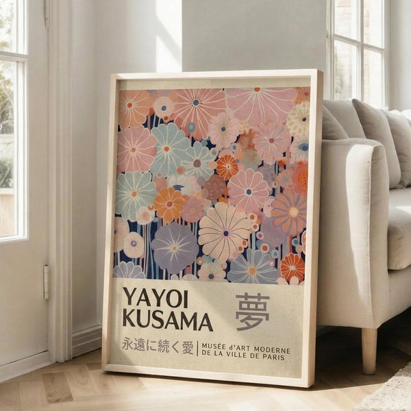 May include: Large framed poster leaning against white wall beside beige sofa in bright living room. Artwork features colorful stylized flowers in pink, orange, blue, and sage green tones with dark stems against cream background. Japanese and French text below floral design. Light wood frame, hardwood floors visible.