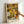 May include: Framed art print displayed against white wall with person's hand visible for scale. Features densely packed floral pattern in warm autumn colors - yellows, oranges, greens, browns, and cream. Abstract flowers and polka dots create overlapping psychedelic design. Text at bottom in Japanese characters and "YAYOI KUSAMA" plus "NAGOYA" in block letters. Light wood frame, approximately poster size.