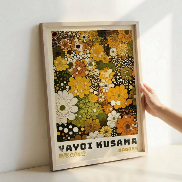May include: Framed art print displayed against white wall with person's hand visible for scale. Features densely packed floral pattern in warm autumn colors - yellows, oranges, greens, browns, and cream. Abstract flowers and polka dots create overlapping psychedelic design. Text at bottom in Japanese characters and "YAYOI KUSAMA" plus "NAGOYA" in block letters. Light wood frame, approximately poster size.