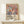 May include: Framed poster with colorful stylized flowers in soft pastels - pink, sage green, orange, and cream blooms with radiating petals against navy background. Text reads "YAYOI KUSAMA" with Japanese characters and "MUSEE D'ART MODERNE VILLE DE PARIS." Natural wood frame sits on light wooden surface next to small white wrapped gift with twine and greenery sprig against neutral wall.