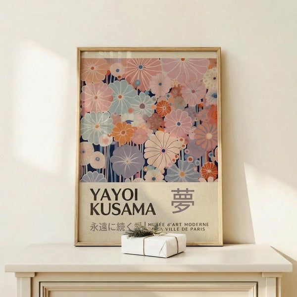 May include: Framed poster with colorful stylized flowers in soft pastels - pink, sage green, orange, and cream blooms with radiating petals against navy background. Text reads "YAYOI KUSAMA" with Japanese characters and "MUSEE D'ART MODERNE VILLE DE PARIS." Natural wood frame sits on light wooden surface next to small white wrapped gift with twine and greenery sprig against neutral wall.