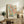 May include: Abstract art poster with colorful organic shapes and dots in pink, teal, yellow and black, featuring text "Yayoi Kusama" and "Musee D'Art Moderne De La Ville De Paris" at bottom. Framed in light wood, displayed on rustic wooden sideboard alongside potted fern, stacked books, ceramic vessels, and brown leather chair in neutral-toned living room.