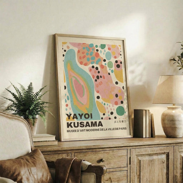 May include: Abstract art poster with colorful organic shapes and dots in pink, teal, yellow and black, featuring text "Yayoi Kusama" and "Musee D'Art Moderne De La Ville De Paris" at bottom. Framed in light wood, displayed on rustic wooden sideboard alongside potted fern, stacked books, ceramic vessels, and brown leather chair in neutral-toned living room.