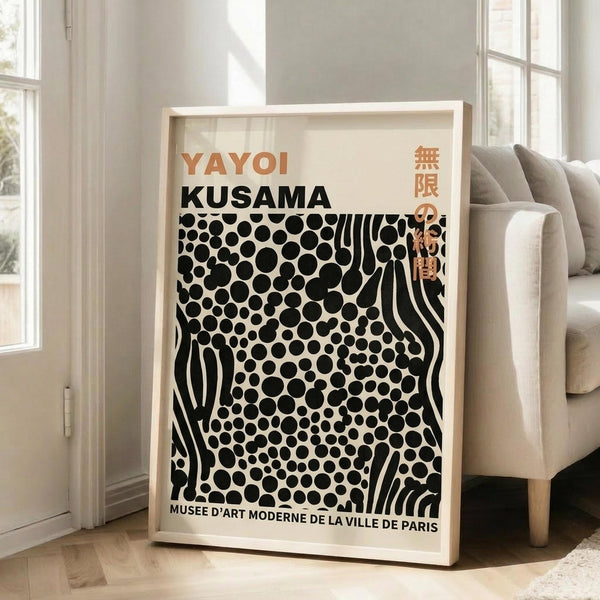 May include: A framed art poster featuring bold black polka dots and flowing striped patterns on cream background, displayed in bright living room corner beside beige sofa. Poster shows orange and black text reading "YAYOI KUSAMA" at top with Japanese characters, and "MUSEE D'ART MODERNE DE LA VILLE DE PARIS" at bottom. Natural wood frame leans against white wall with tall windows casting soft shadows.